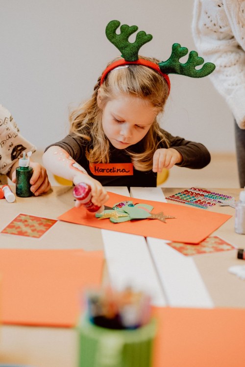 A girl in plush reindeer ears paints a Christmas tree cut out of paper with glitter from a tube